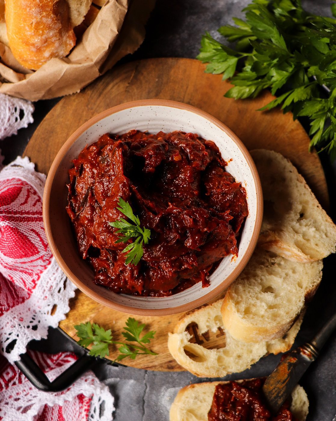 Bowl of ajvar Balkan pepper spread garnished with parsley, served on a wooden board with sliced rustic bread, fresh herbs, and a red patterned cloth in the background.