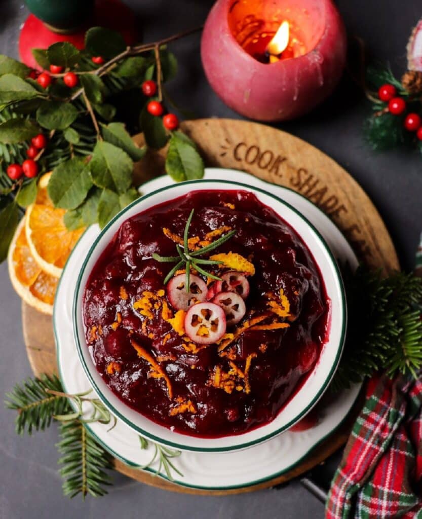 A bowl of festive amaretto cranberry sauce topped with orange zest, sliced cranberries, and rosemary, surrounded by holiday greenery and dried orange slices.