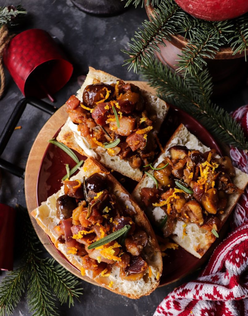Close-up of Festive Chestnut Pancetta Bruschetta topped with caramelized onions, chestnuts, and orange zest on toasted bread, served on a red plate with holiday greenery and candles in the background.