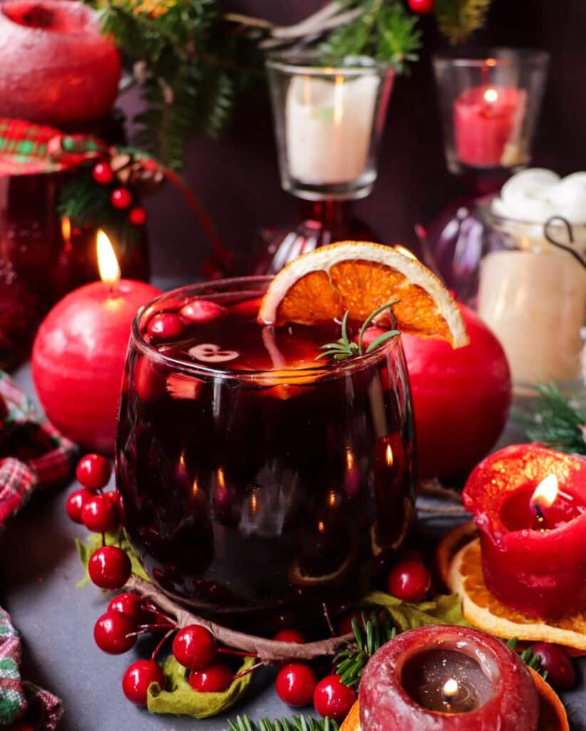 A festive glass of cranberry amaretto sangria garnished with dried orange slices, fresh cranberries, and rosemary, surrounded by glowing red candles and holiday décor.