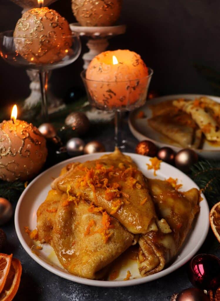 A festive plate of Orange Liqueur Crêpes Suzette drizzled with buttery orange sauce and garnished with orange zest, surrounded by glowing candles, baubles, and evergreen branches for a warm holiday setting.