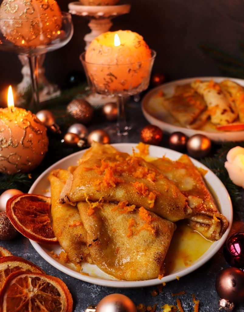 A festive plate of Orange Liqueur Crêpes Suzette drizzled with buttery orange sauce and garnished with orange zest, surrounded by glowing candles, baubles, and evergreen branches for a warm holiday setting.