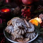 Plate of festive spiced shortbread cookies for Santa dusted with powdered sugar, arranged with dried orange slices, cinnamon sticks, and star anise, surrounded by evergreen branches and candlelight.