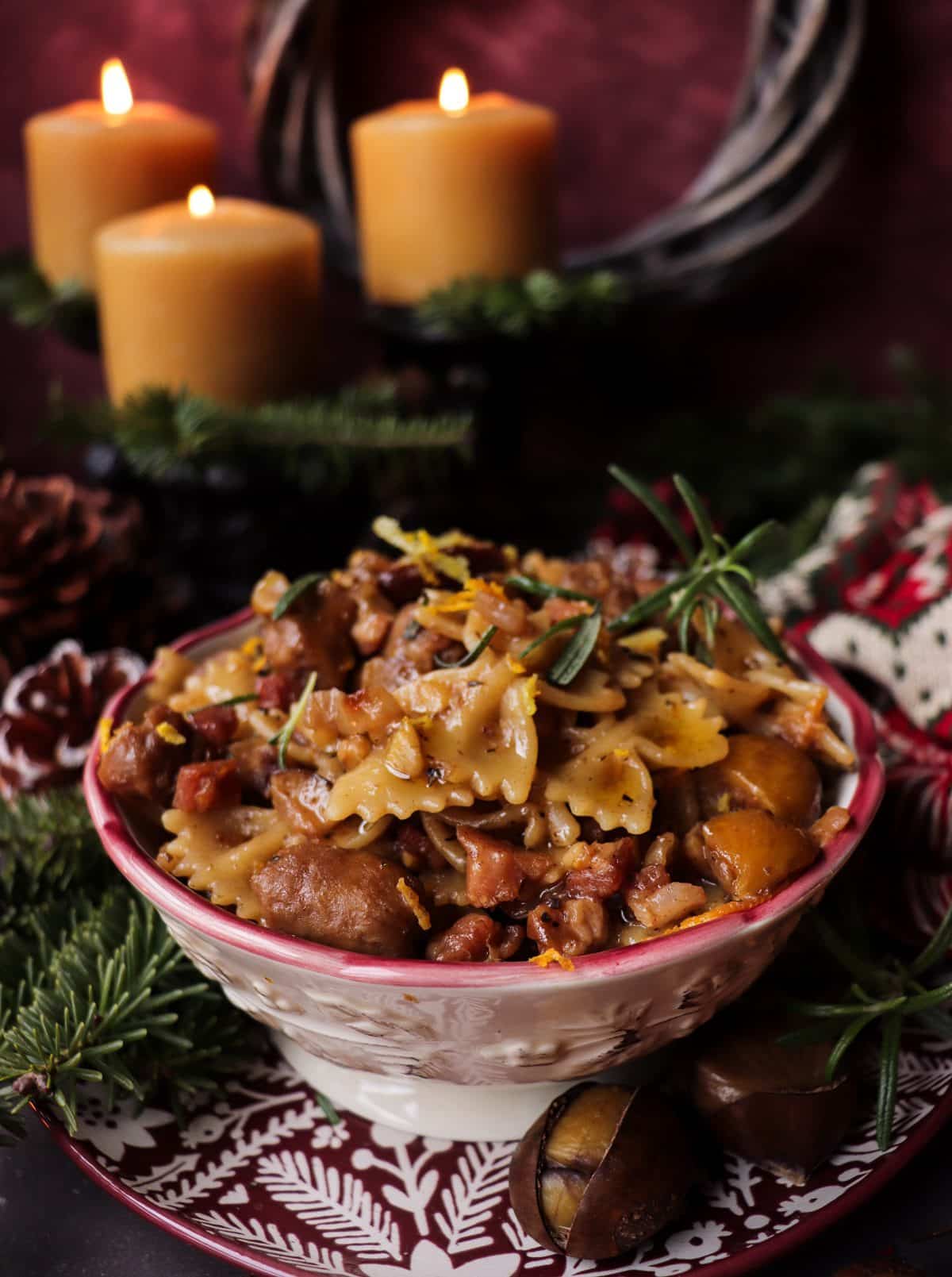 Bowl of festive chestnut sausage pasta with pancetta, rosemary, and orange zest, served on a rustic plate with a fork and glass of brandy.