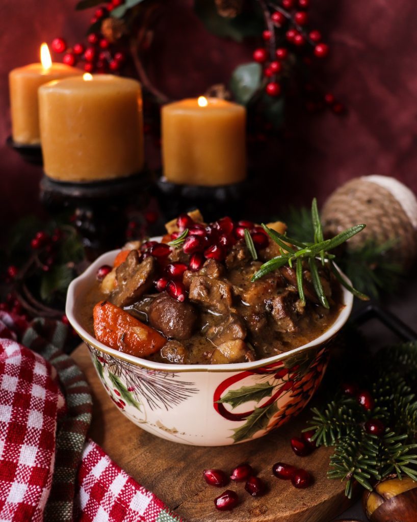 A festive bowl of venison stew with carrots, mushrooms, chestnuts, and pancetta, topped with fresh pomegranate seeds and rosemary. Served in a Christmas-patterned dish surrounded by candles and evergreens, creating a warm holiday atmosphere.