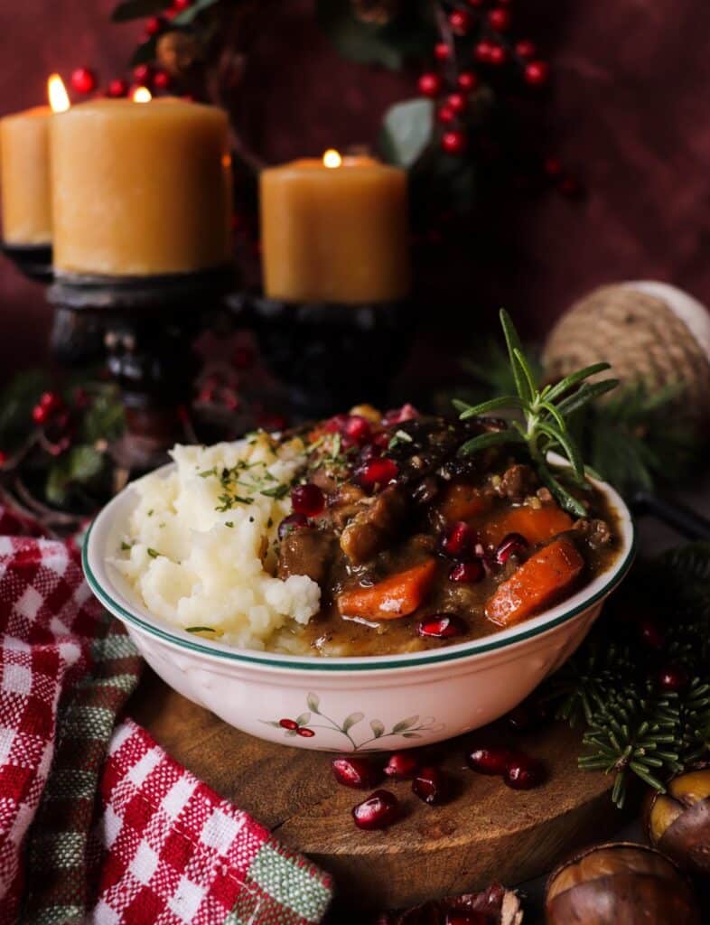 A hearty serving of festive venison stew with chestnuts, carrots, and pancetta, topped with pomegranate seeds and fresh rosemary, served alongside creamy mashed potatoes. A warm, rustic holiday meal surrounded by candles and evergreens.