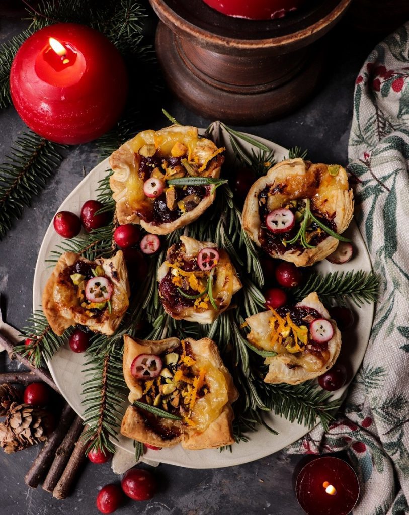 A festive plate of cranberry brie puff pastry bites topped with pistachios, rosemary, and orange zest, surrounded by fresh cranberries and evergreen sprigs — a perfect holiday appetiser.