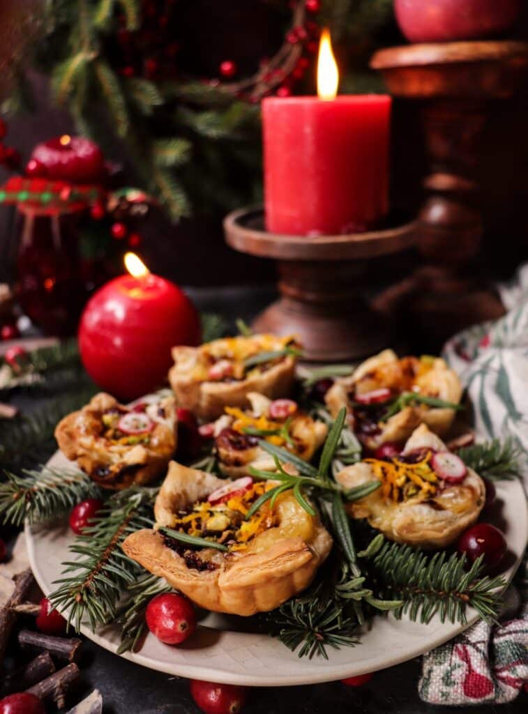 Golden cranberry brie puff pastry bites topped with pistachios, rosemary, and orange zest, surrounded by evergreen sprigs and festive candlelight — a perfect holiday appetizer.