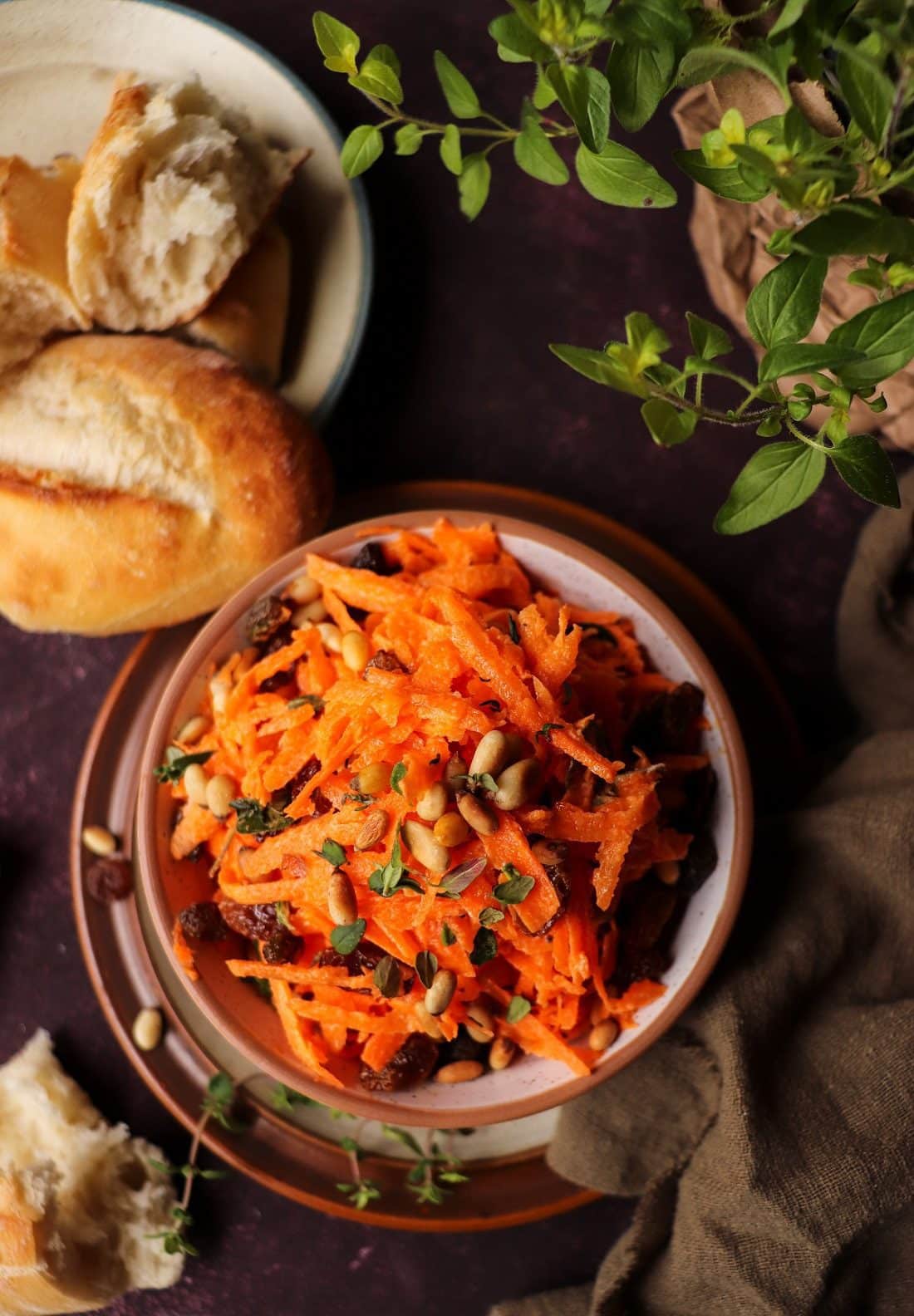 Carrot salad with pine nuts and raisins served in a rustic ceramic bowl, garnished with fresh thyme and toasted pine nuts, with bread and herbs in a warm, rustic background.