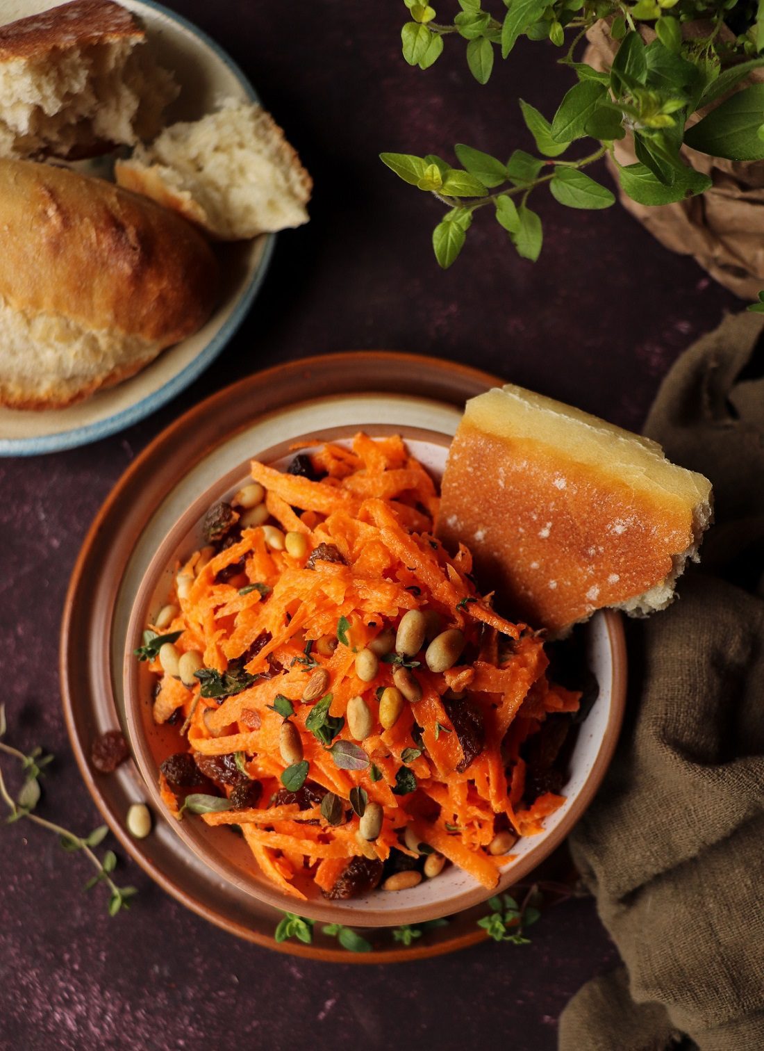 Carrot salad with pine nuts and raisins served in a rustic ceramic bowl, garnished with fresh thyme and toasted pine nuts, with bread and herbs in a warm, rustic background.