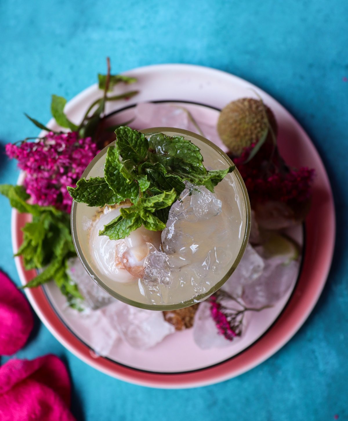 Top view of a refreshing lychee mojito with ice, mint leaves, and juicy lychees, photographed on a soft pink plate with scattered ice and bright turquoise backdrop.