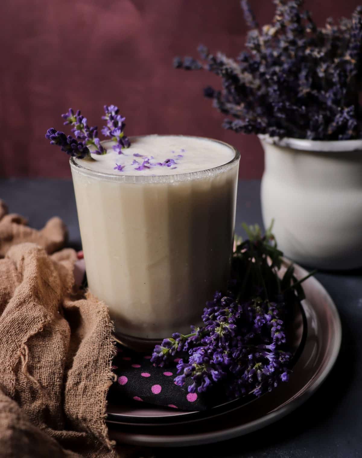 Lavender milkshake in a clear glass, garnished with fresh lavender blossoms and served on a plate with a napkin and lavender sprigs.