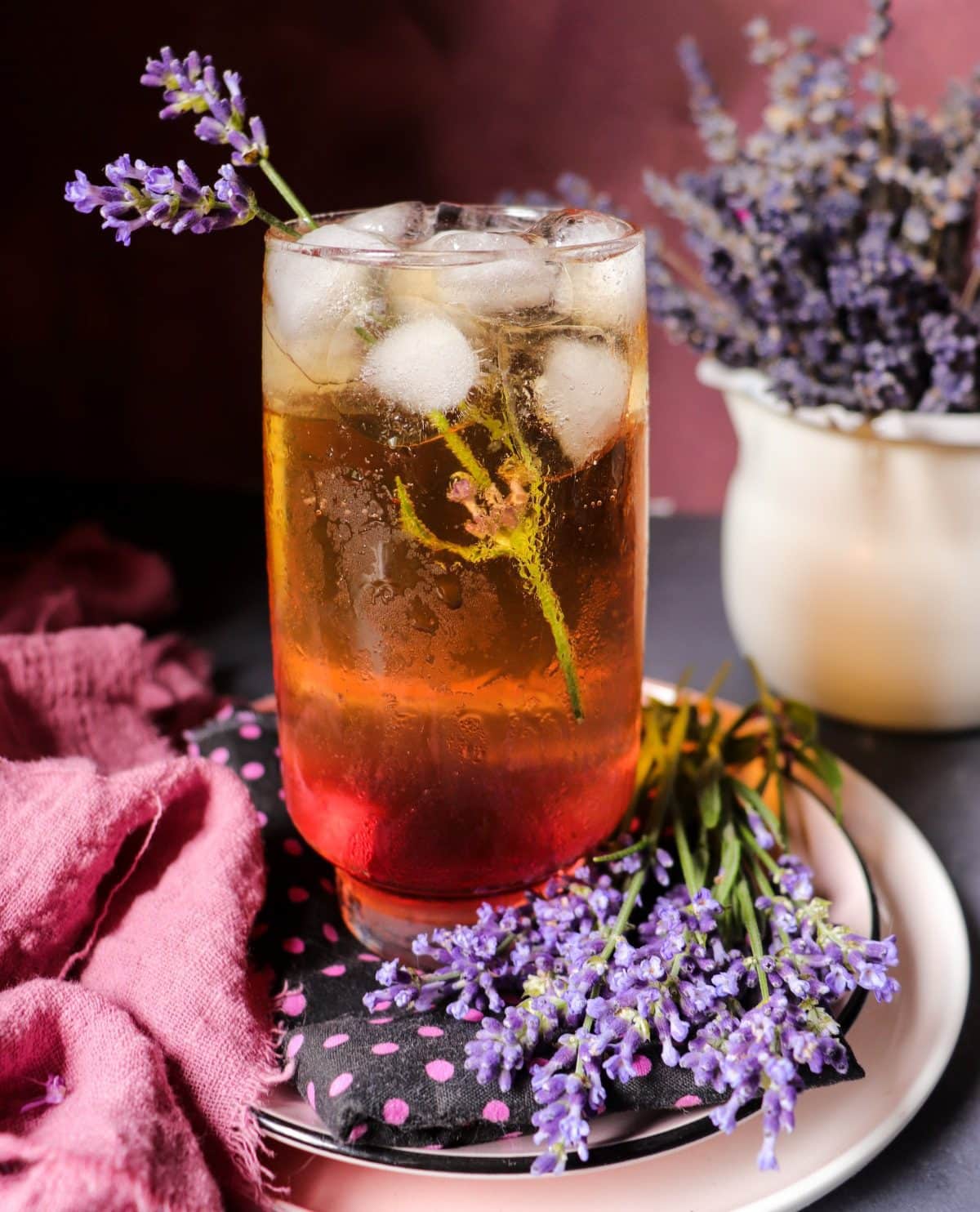 Lavender apple cider cocktail served over ice in a tall glass, garnished with a lavender sprig and surrounded by fresh lavender on a plate, with soft purple tones and a rustic background.
