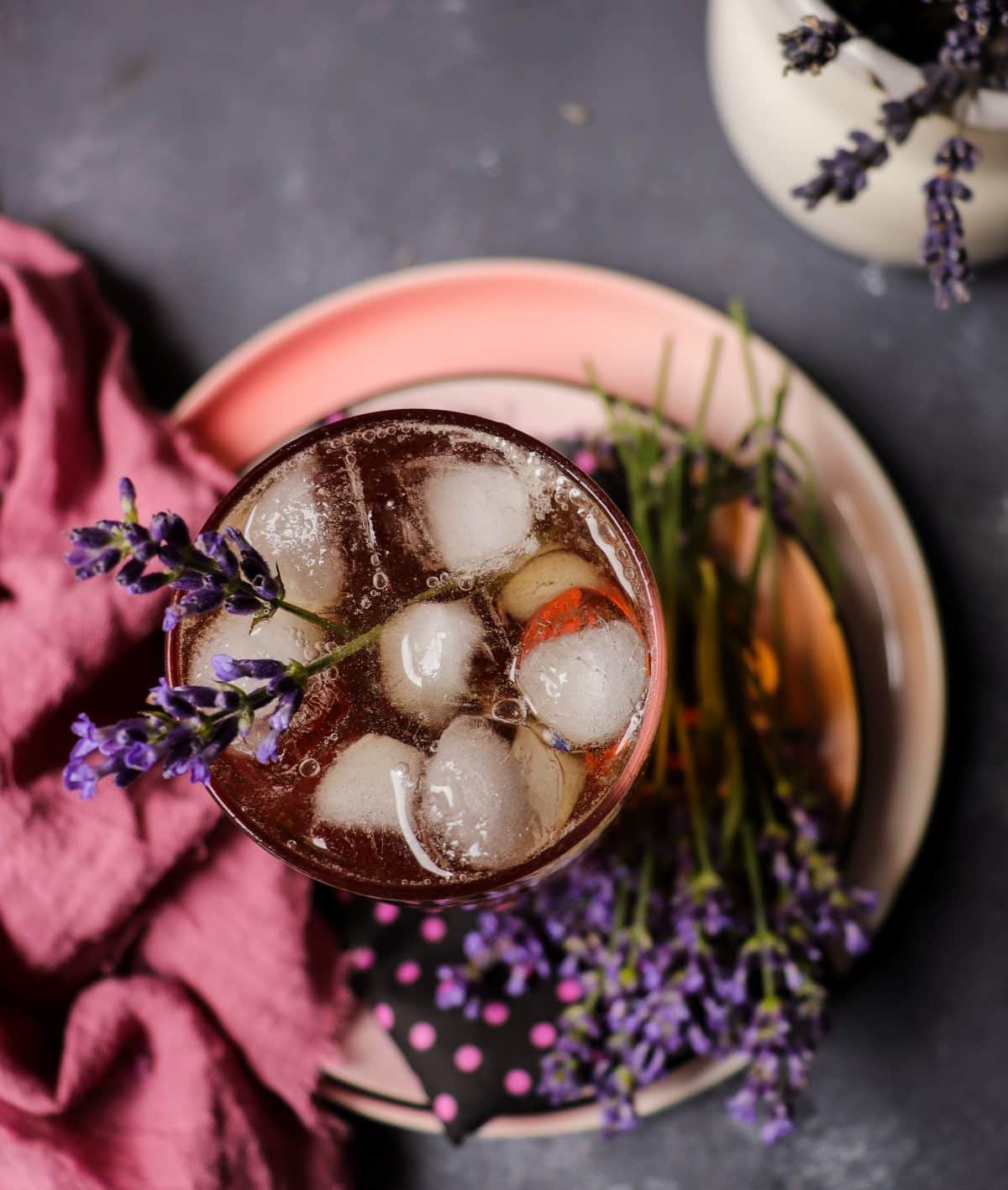Lavender apple cider cocktail served over ice in a tall glass, garnished with a lavender sprig and surrounded by fresh lavender on a plate, with soft purple tones and a rustic background.