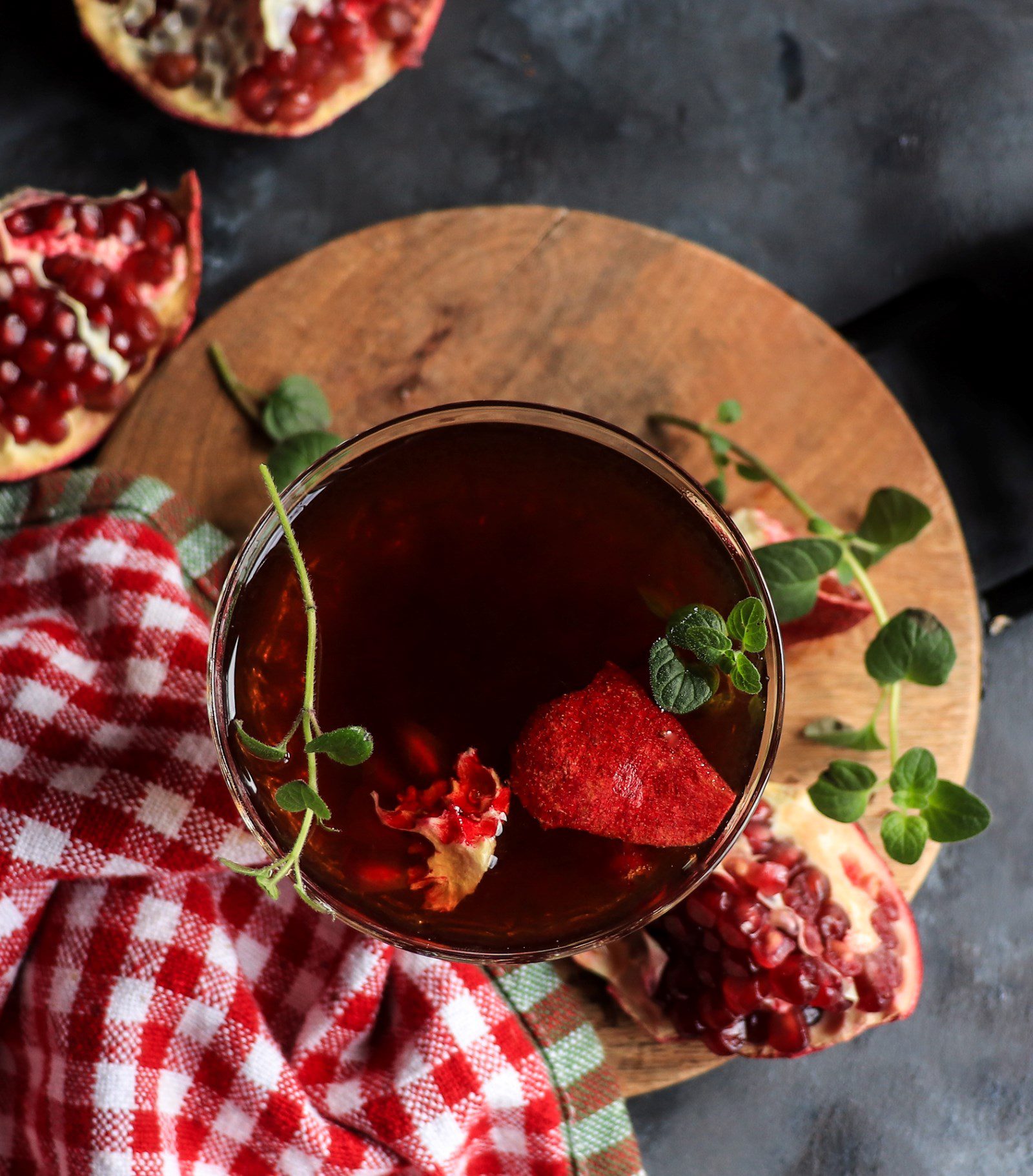 Top view of a pomegranate brandy cocktail with rich red tones, pomegranate garnish, and fresh herbs on a rustic board.