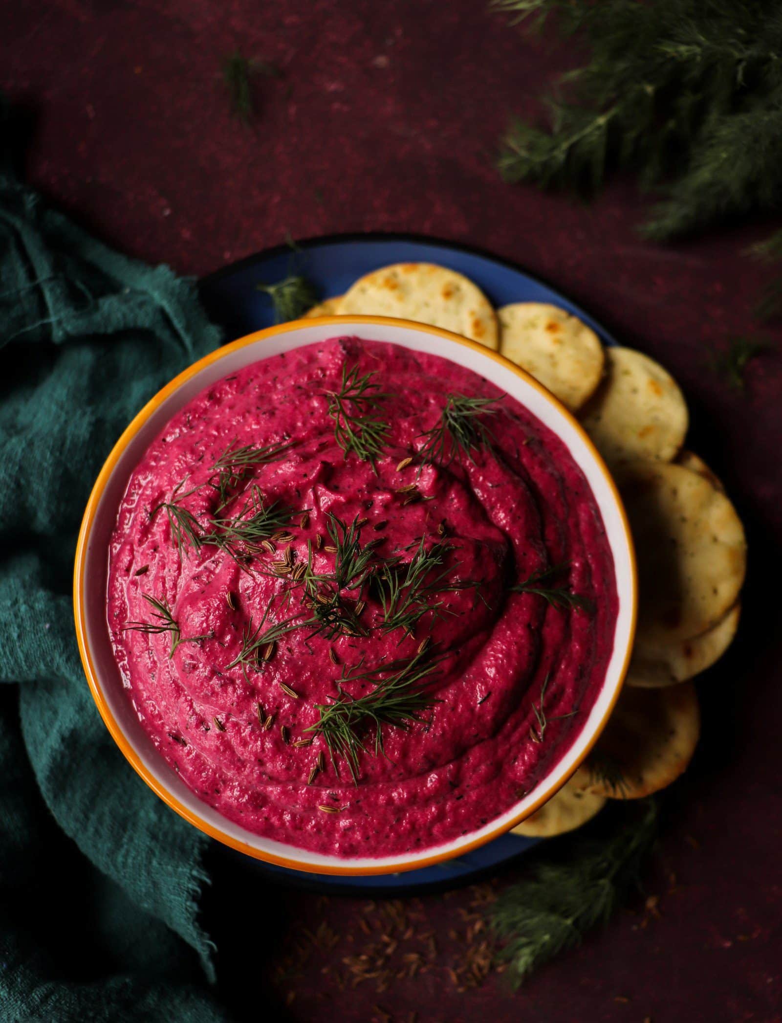 Beetroot Dip with Caraway Seeds and Dill served in a bowl and garnished with fresh dill and caraway seeds, surrounded by crackers on a dark red background.