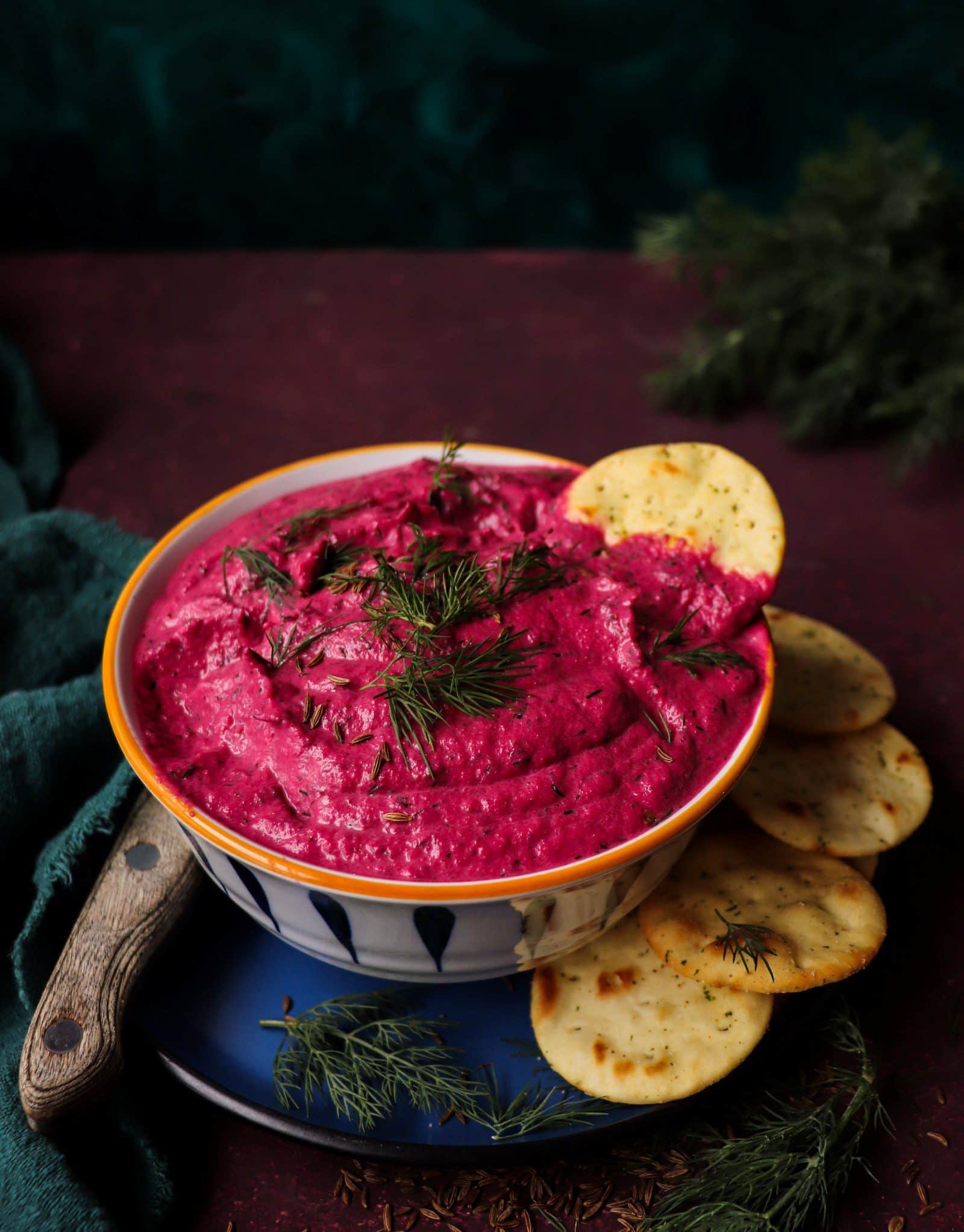 Beetroot Dip with Caraway Seeds and Dill served in a bowl and garnished with fresh dill and caraway seeds, surrounded by crackers on a dark red background.