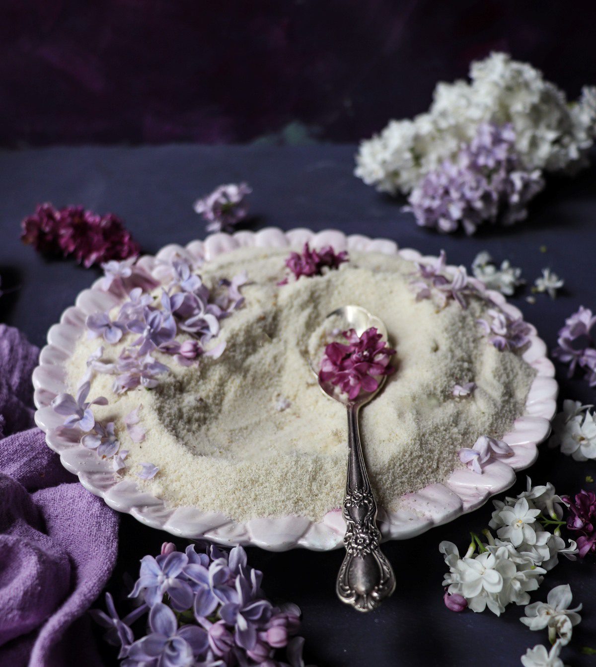 Lilac sugar spread on a plate with fresh lilac blossoms and a spoonful of infused sugar, showing the delicate floral sugar texture and soft purple petals.