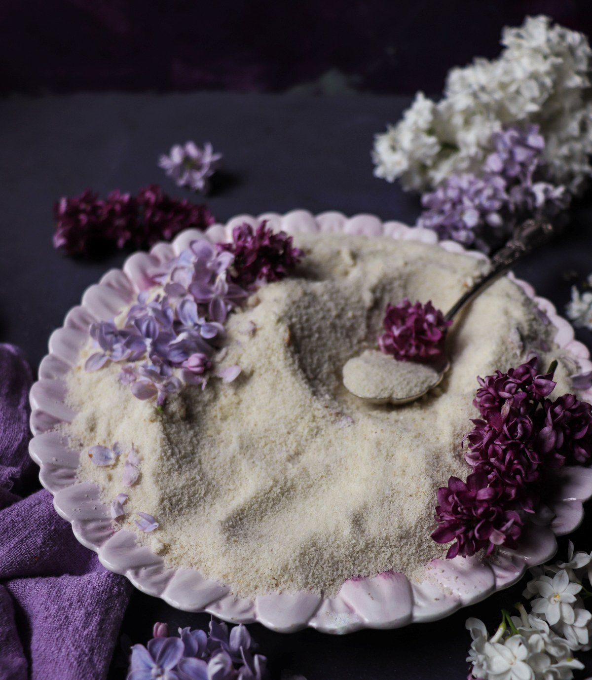 Lilac sugar spread on a plate with fresh lilac blossoms and a spoonful of infused sugar, showing the delicate floral sugar texture and soft purple petals.
