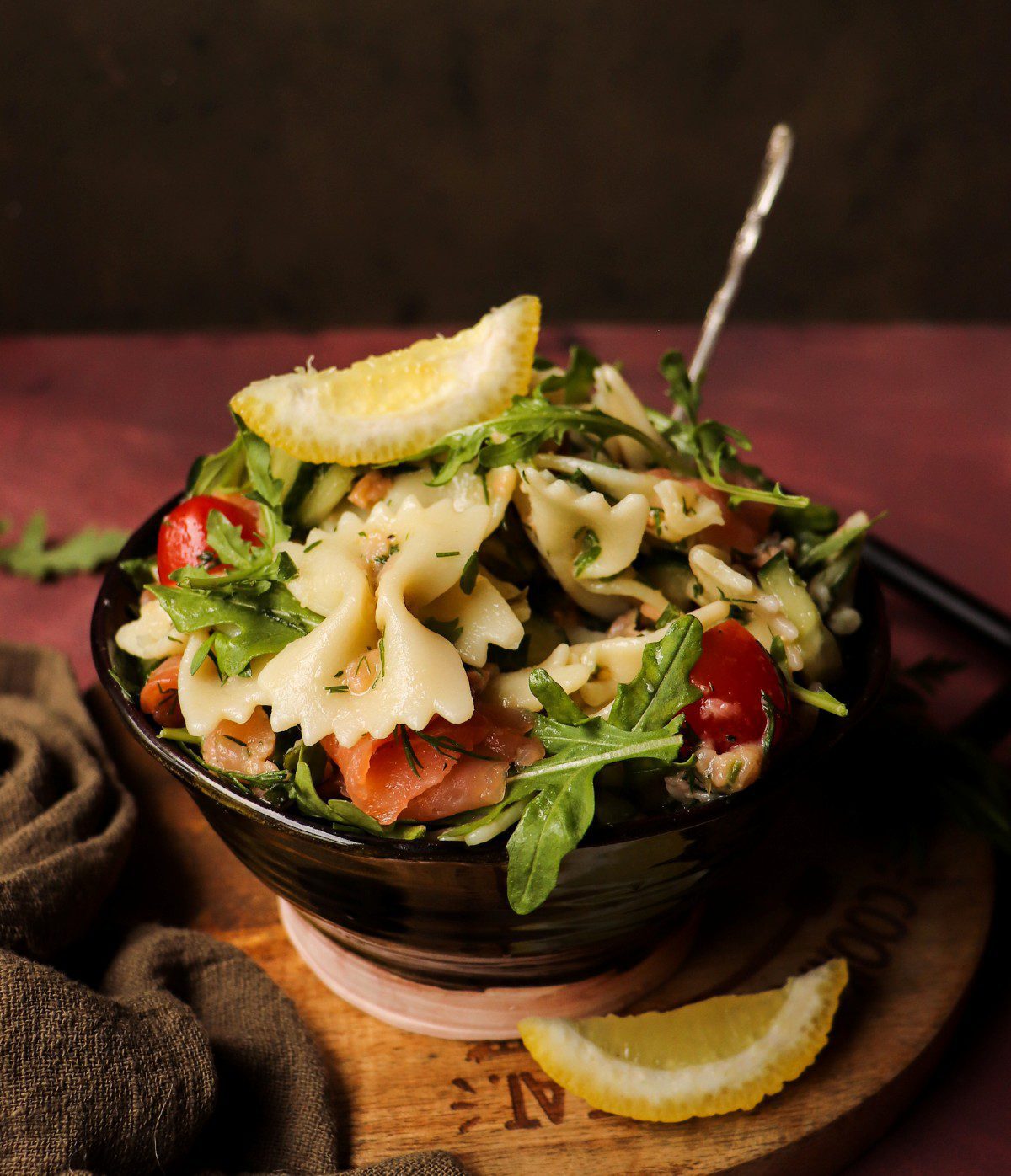 Smoked salmon pasta salad with arugula, cherry tomatoes and cucumber in a bowl, served with a lemon wedge on the side.