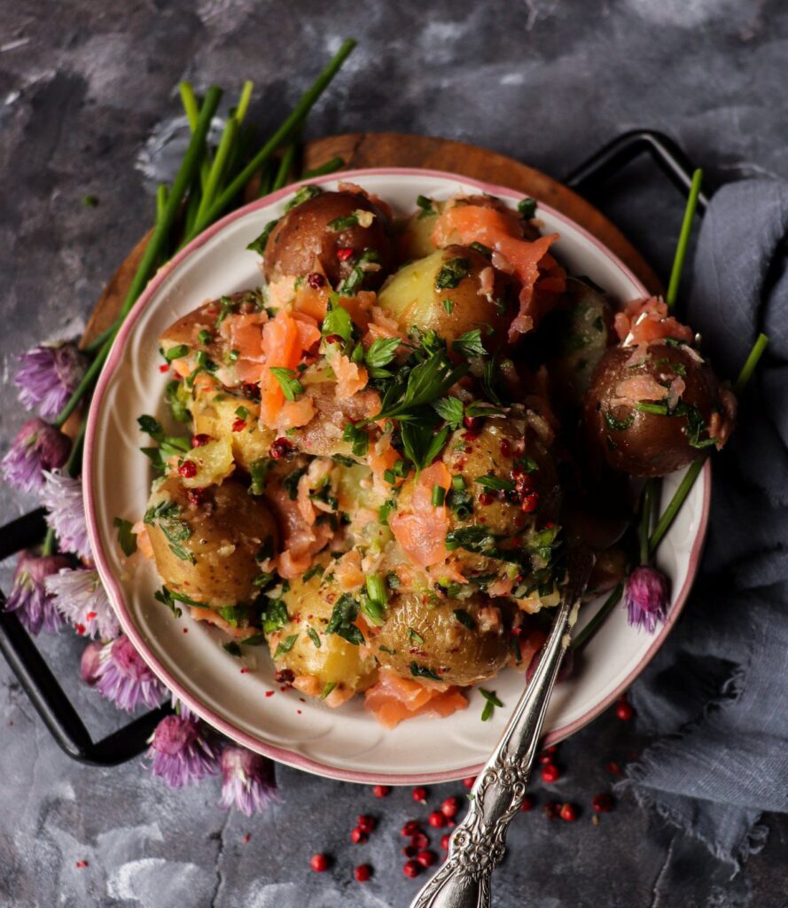 Smoked salmon potato salad with baby potatoes, fresh herbs, chopped chives, capers, and crushed pink peppercorns, dressed lightly with olive oil and served in a shallow bowl.