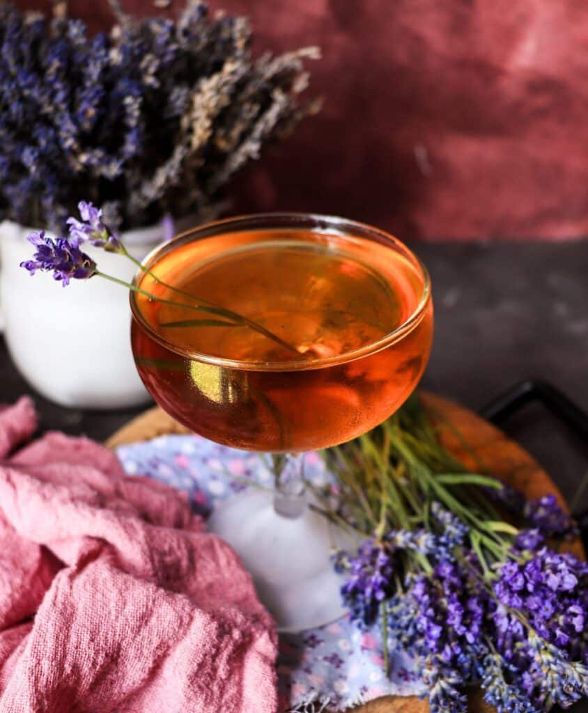 Apricot lavender cocktail with gin and apricot brandy in a coupe glass, garnished with fresh lavender, styled with dried lavender bundles and a soft pink linen backdrop.