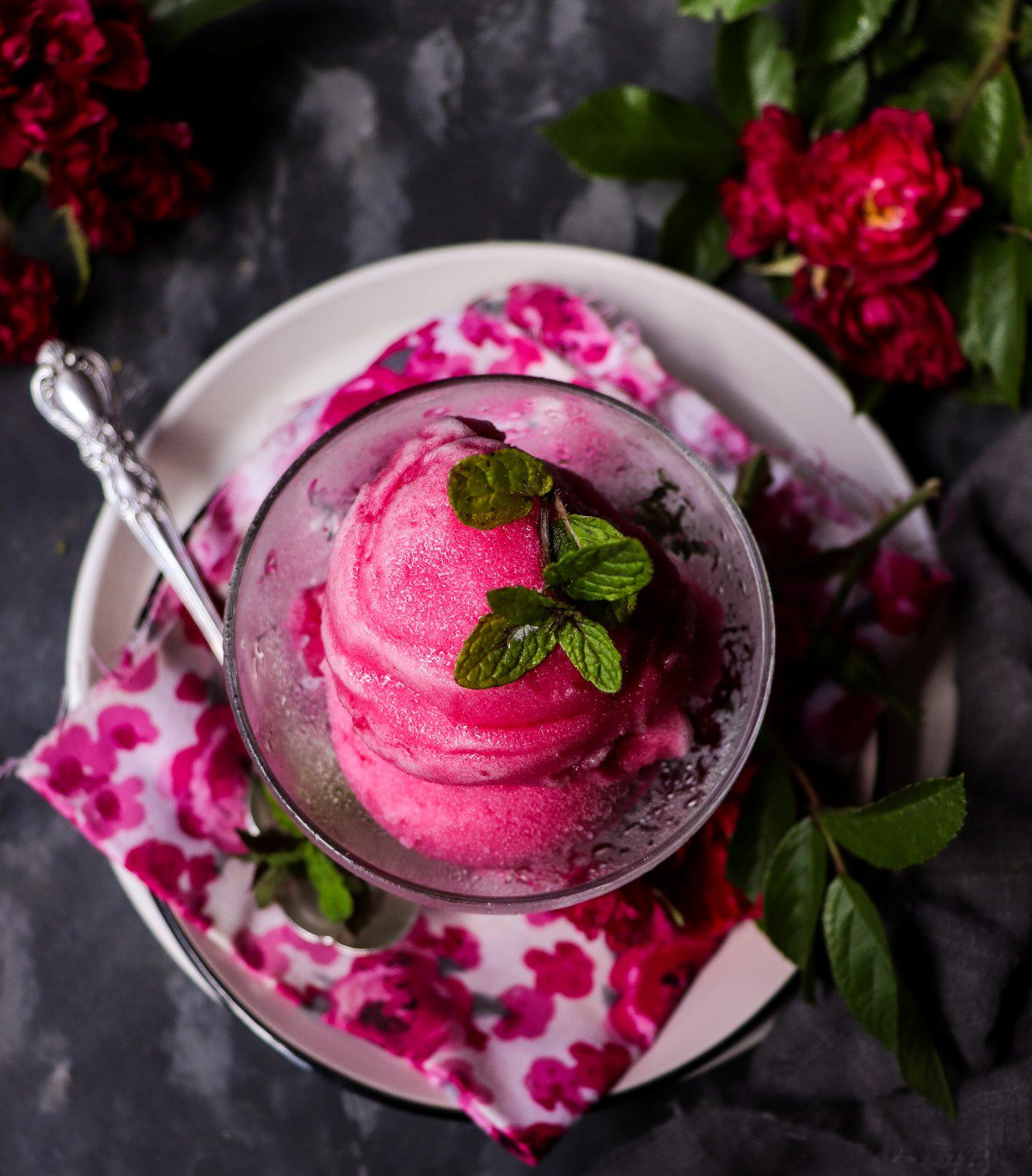 Rose sorbet served in a glass dessert bowl with a mint garnish, surrounded by fresh red roses.