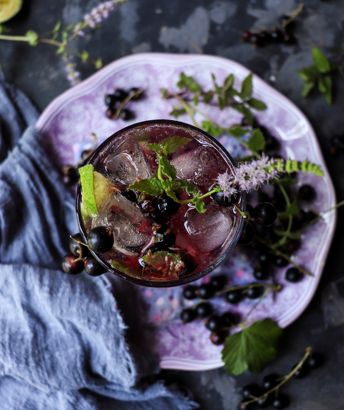 Top view of a blackcurrant mojito with ice, mint leaves, lime wedges, and blackcurrants, highlighting its rich berry colour.