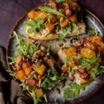 Roasted Butternut Squash Bruschetta topped with crispy ham, sage, goat cheese, and arugula on toasted bread slices — a warm and cozy fall appetizer.