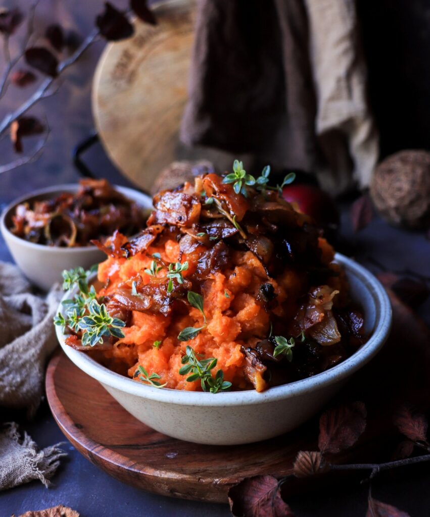 Bowl of creamy savory sweet potato mash topped with golden caramelized onions, crisp prosciutto, and fresh thyme sprigs — a rich and festive side dish perfect for holiday dinners.