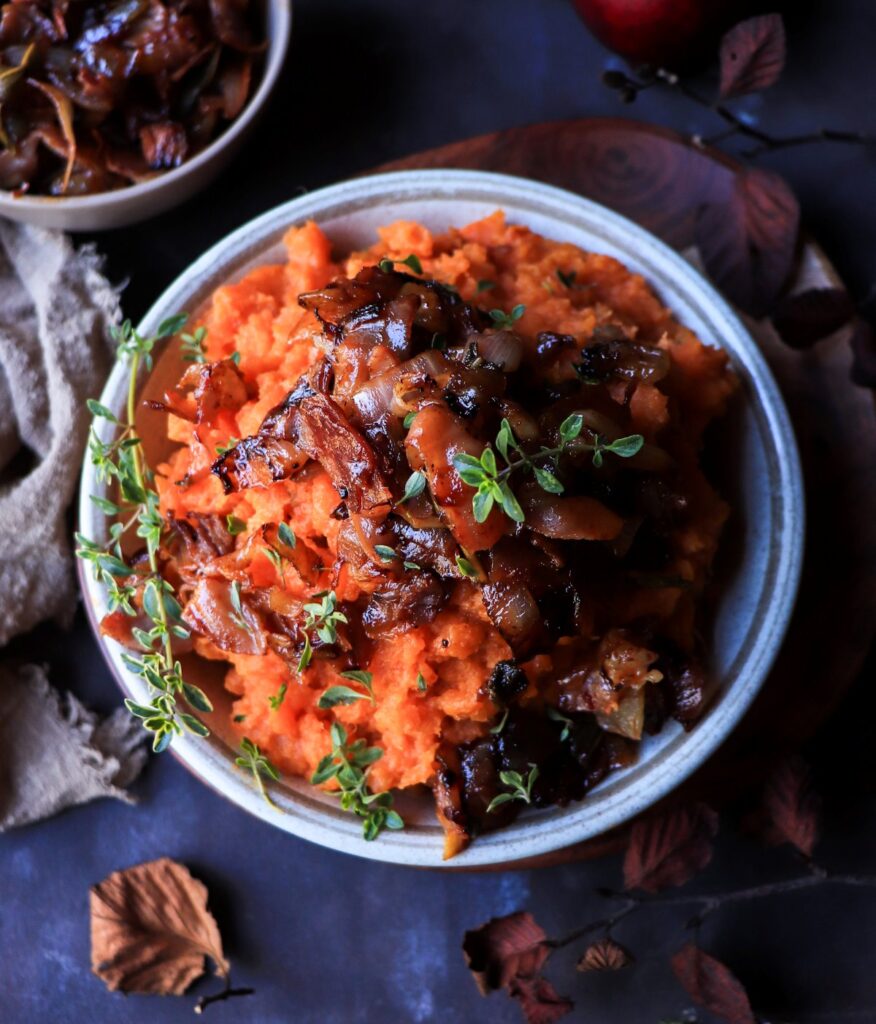 Overhead view of savory sweet potato mash topped with golden caramelized onions, crispy prosciutto, and sprigs of thyme — a warm, festive side dish perfect for holiday dinners.