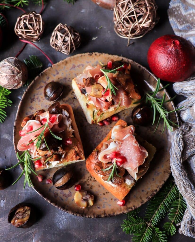 Overhead view of caramelized onion and chestnut bruschetta with prosciutto, rosemary, and pomegranate on a rustic plate surrounded by chestnuts and festive greenery.