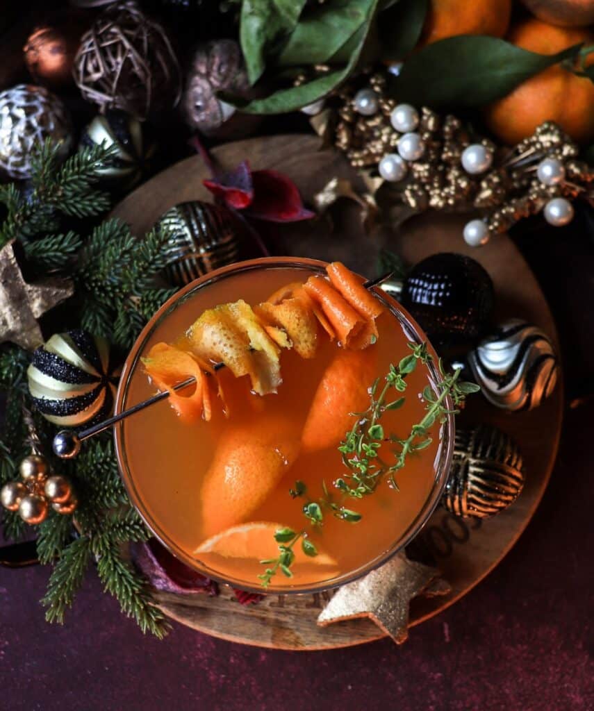 Top-down view of a clementine honey brandy cocktail with clementine peel garnish and fresh thyme, set among Christmas greenery and shimmering holiday decorations.