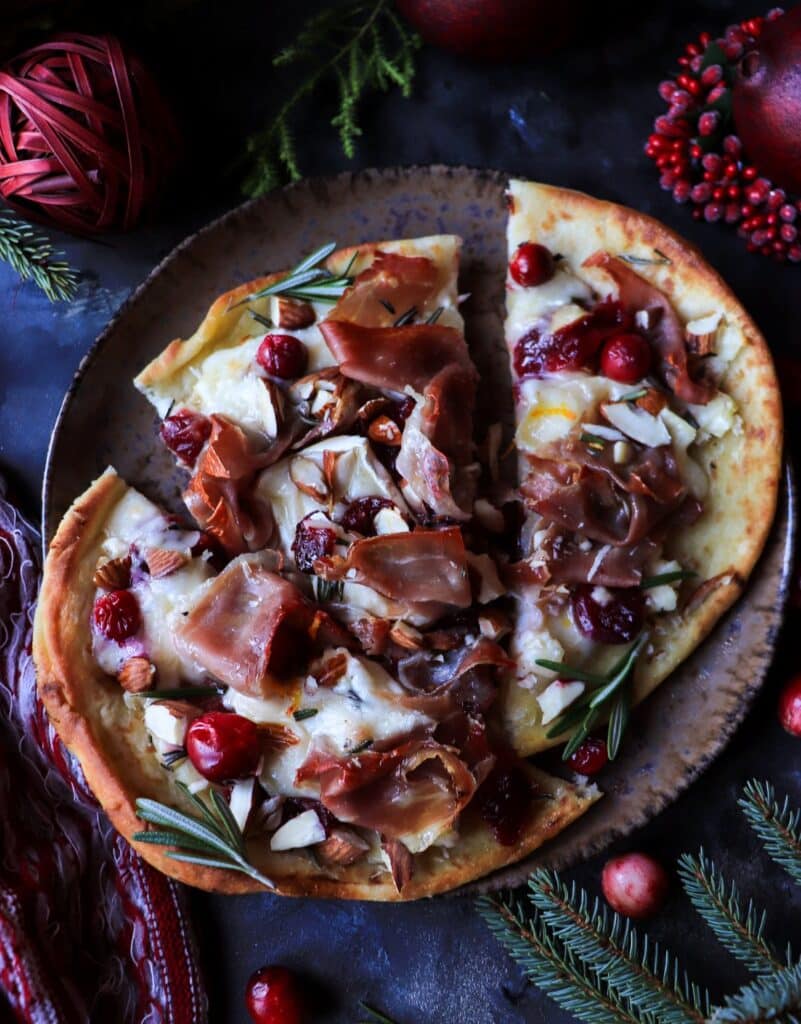 Close-up of melted brie and prosciutto on cranberry flatbread, garnished with almonds and rosemary for an elegant Christmas appetizer.