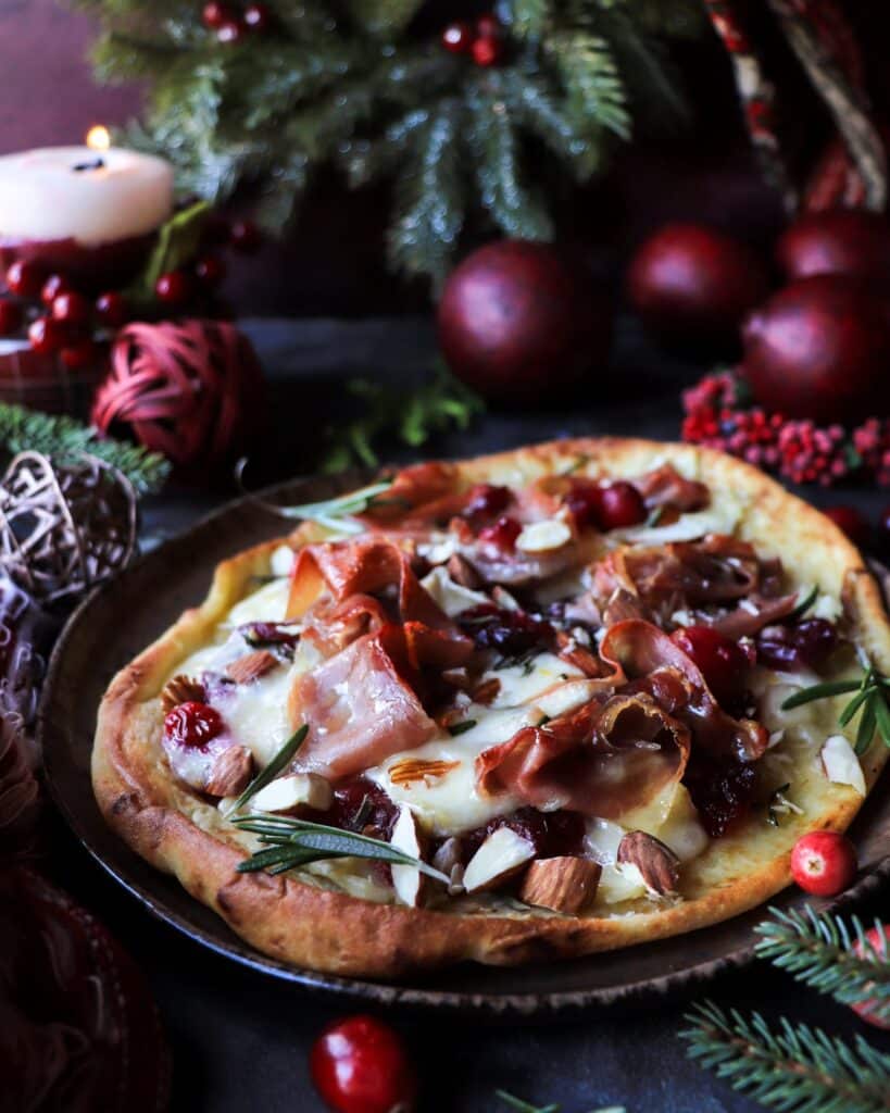 Festive cranberry brie prosciutto flatbread on naan bread, topped with melted brie, prosciutto, and rosemary, styled with candles and evergreens for a cozy holiday table.
