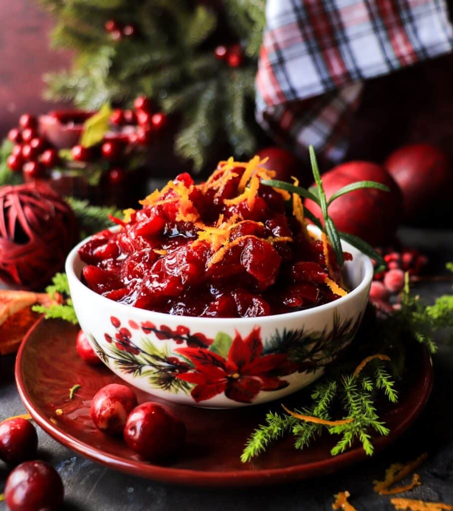 Side view of glossy cranberry maple orange sauce garnished with orange zest and rosemary, styled with Christmas decor and warm candlelight.