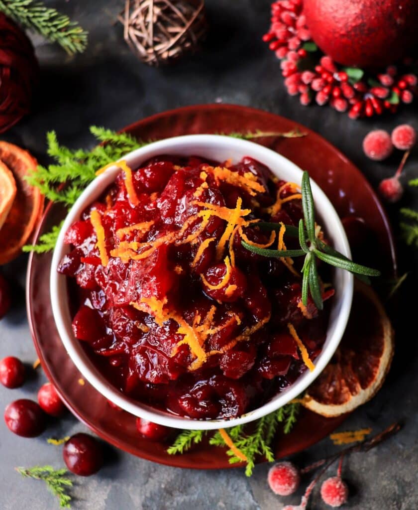 Cranberry maple orange sauce in a festive bowl, topped with fresh orange zest and a sprig of rosemary, surrounded by holiday greenery.