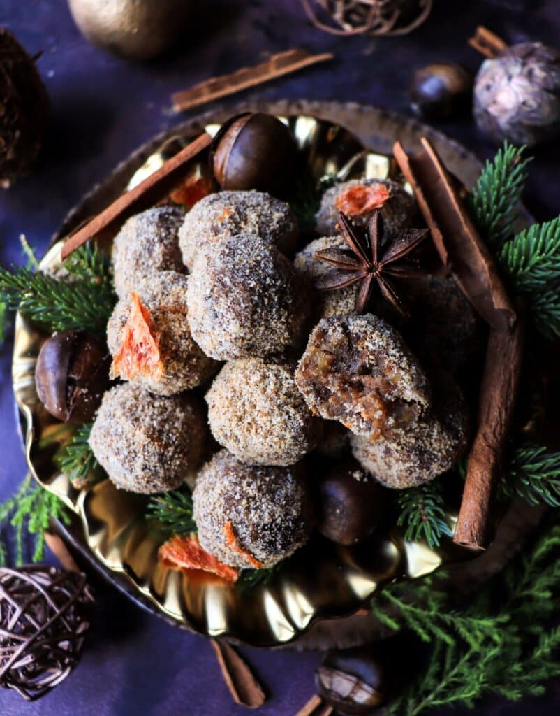 Date chestnut bliss ball cut open to show the soft, spiced interior, surrounded by whole bliss balls coated in toasted almond meal with orange zest, cinnamon, cloves, chestnuts, and evergreen sprigs.