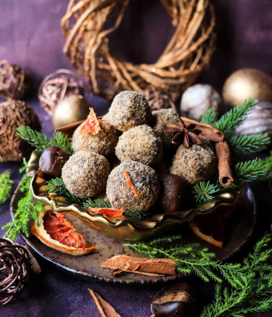 Date chestnut bliss balls coated in toasted almond meal, arranged on a gold scalloped dish with fresh evergreens, dried orange slices, cinnamon sticks, and chestnuts.