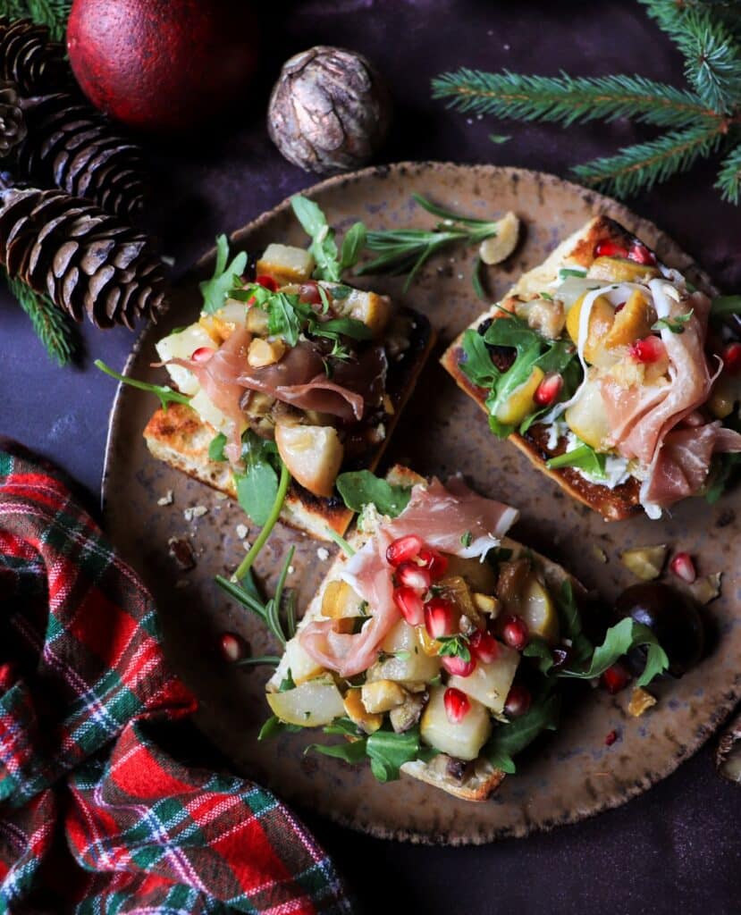 Festive pear chestnut bruschetta with prosciutto, arugula, and pomegranate on rustic plate surrounded by pinecones and holiday decor.