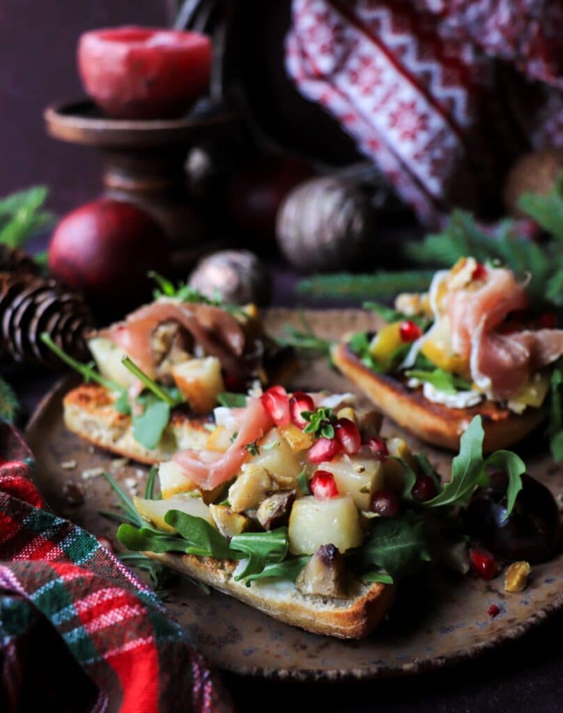 Close-up of pear chestnut bruschetta topped with caramelized pears, chestnuts, arugula, and pomegranate, set against cozy Christmas background.