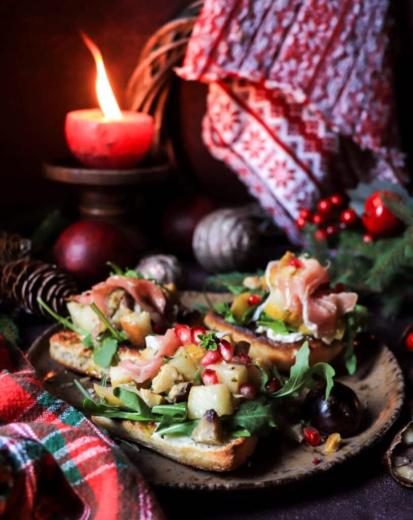 Festive pear chestnut bruschetta with caramelized pears, chestnuts, arugula, and prosciutto, styled with holiday candle and red plaid cloth.