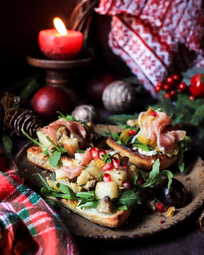 Close-up of festive pear chestnut bruschetta with prosciutto and pomegranate on toasted bread, styled with candlelight and Christmas decorations.