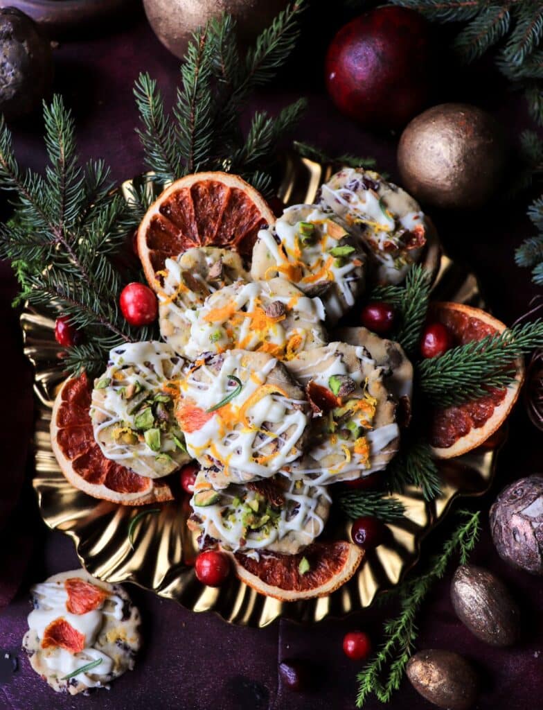 Close top-down shot of shortbread cookies with pistachios, cranberries, orange zest, and white chocolate drizzle, styled with dried citrus and fresh greenery