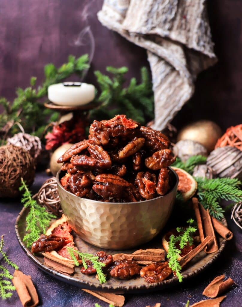 A festive bowl of glossy toffee orange brandy pecans surrounded by cinnamon sticks, dried orange slices, and evergreen branches — rich stovetop caramelized nuts perfect for the holidays.