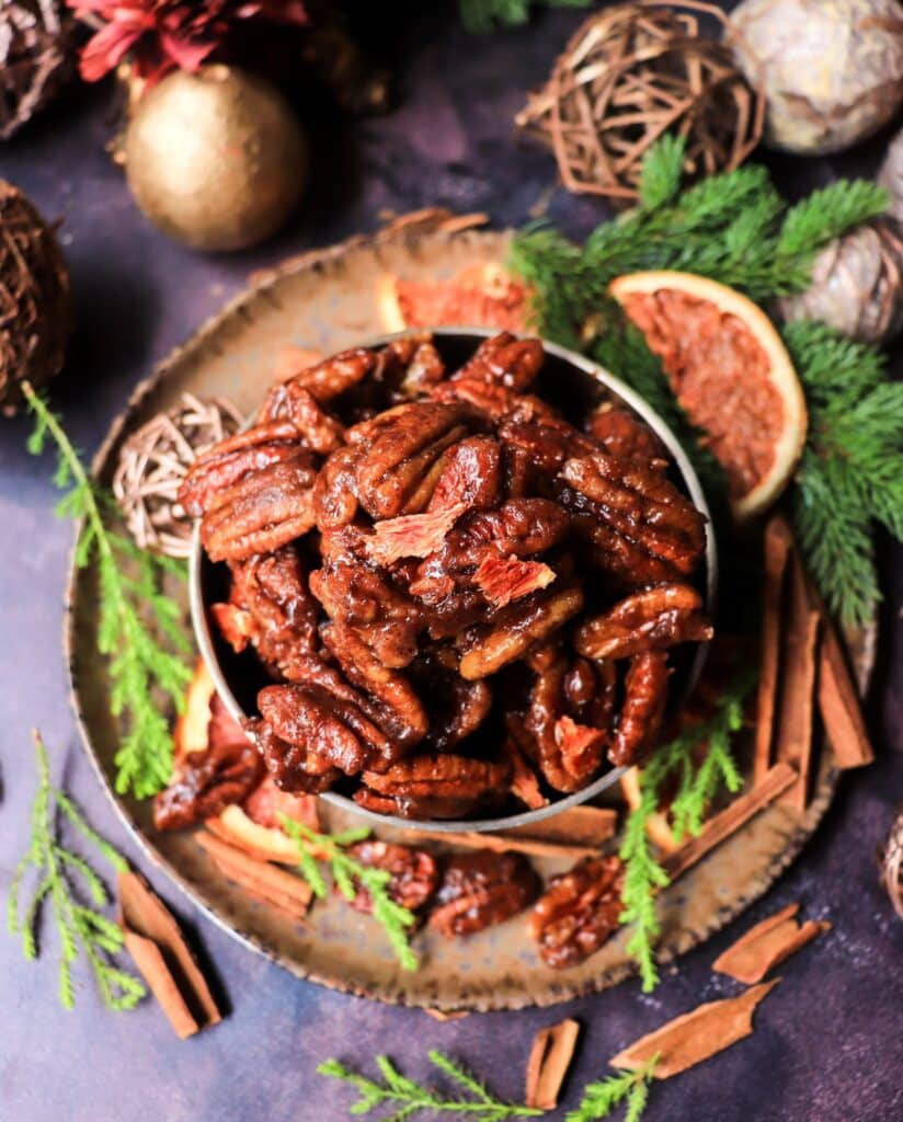 A festive bowl of stovetop toffee orange brandy pecans shot from above, surrounded by cinnamon sticks, evergreen branches, and dried orange slices on a rustic plate.