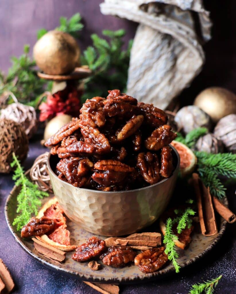 A festive bowl of glossy toffee orange brandy pecans surrounded by cinnamon sticks, golden decor, dried orange slices, and evergreen branches — rich stovetop caramelized nuts perfect for the holidays.