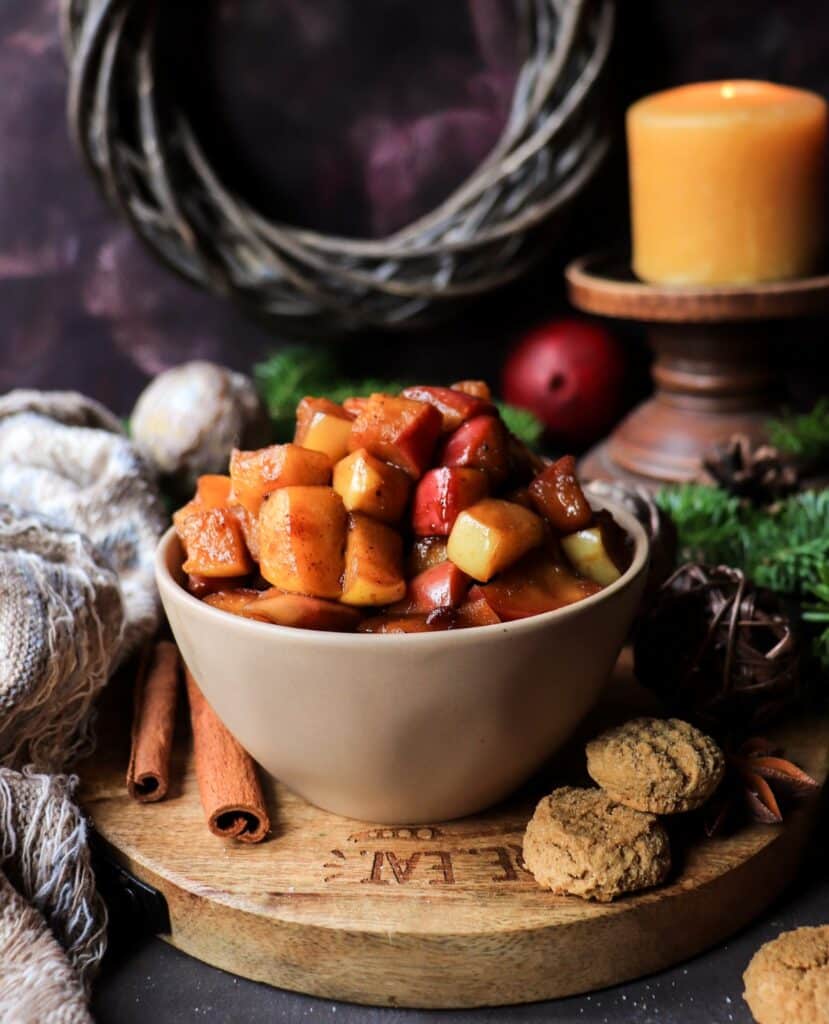 Gingerbread Caramelized Apples in a rustic bowl surrounded by cozy textile, evergreens, candles, cinnamon sticks, star anise, and gingersnaps creating a cozy, festive atmosphere.