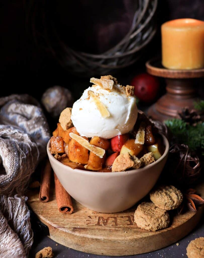 Gingerbread Caramelized Apples in a rustic bowl, garnished with ice cream, caramelized ginger, and crushed ginger cookies,  surrounded by cozy textile, evergreens, candles, cinnamon sticks, star anise, and gingersnaps creating a cozy, festive atmosphere.