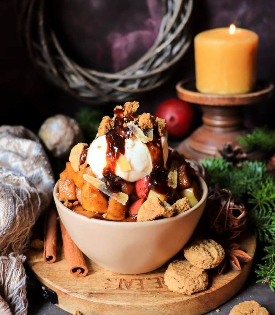 Gingerbread Caramelized Apples in a rustic bowl, garnished with ice cream, caramelized ginger, and crushed ginger cookies,  surrounded by cozy textile, evergreens, candles, cinnamon sticks, star anise, and gingersnaps creating a cozy, festive atmosphere.